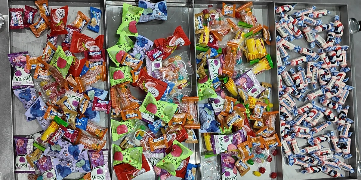 Various candies in their original packaging—including Skittles, gummy corn, and White Rabbit creamy candies—are arranged on multiple stainless steel freeze-drying trays, ready to be selected and tested by the market.