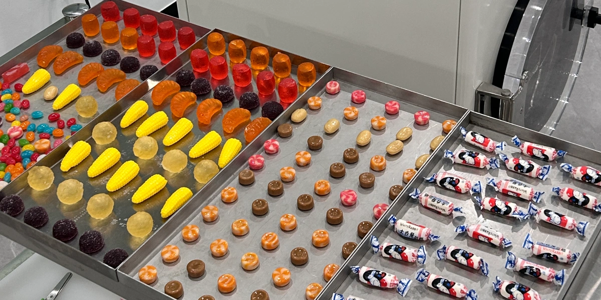 Various types of candies—including Skittles, gummy corn, and White Rabbit creamy candies—are neatly arranged on multiple stainless steel freeze-drying trays, ready to be loaded into the freeze-dryer.